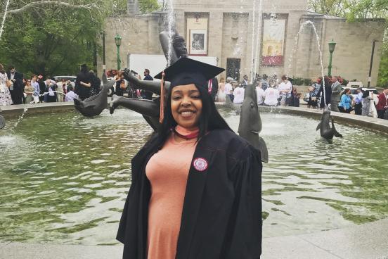 Smiling woman in a graduation robe standing in front of a fountain.