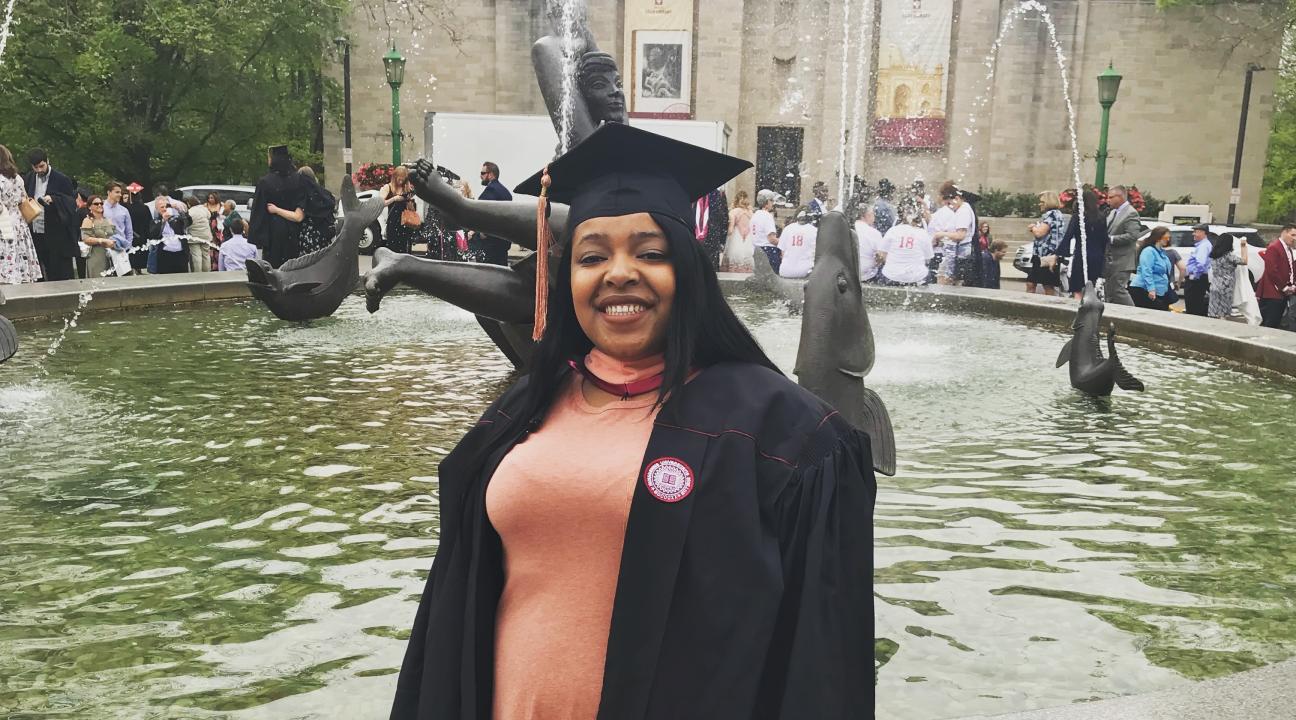 Smiling woman in a graduation robe standing in front of a fountain.