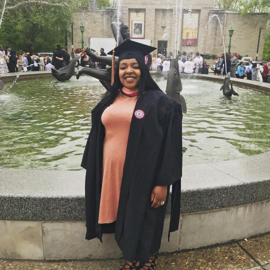 Smiling woman in a graduation robe standing in front of a fountain.